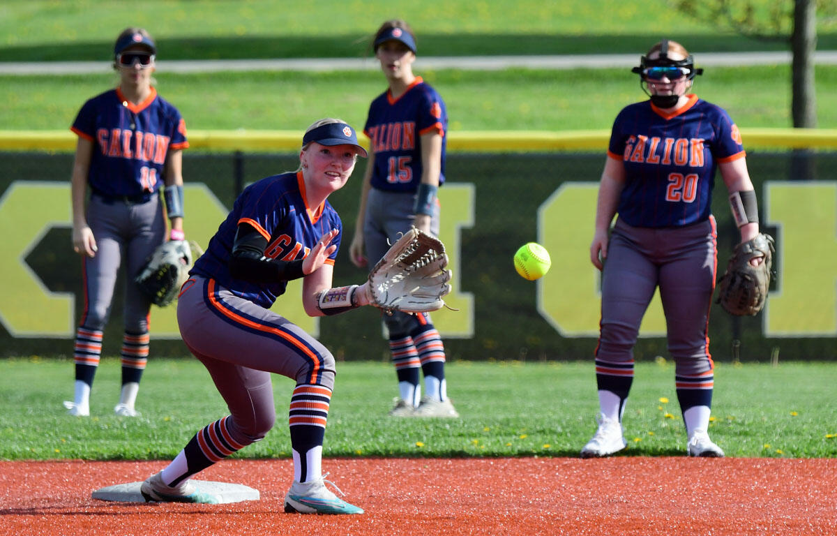 Galion softball player in navy and orange reaches with her glove to catch a bright ball near a base, teammates watching in the outfield.