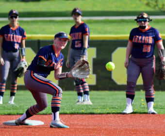 Galion softball player in navy and orange reaches with her glove to catch a bright ball near a base, teammates watching in the outfield.