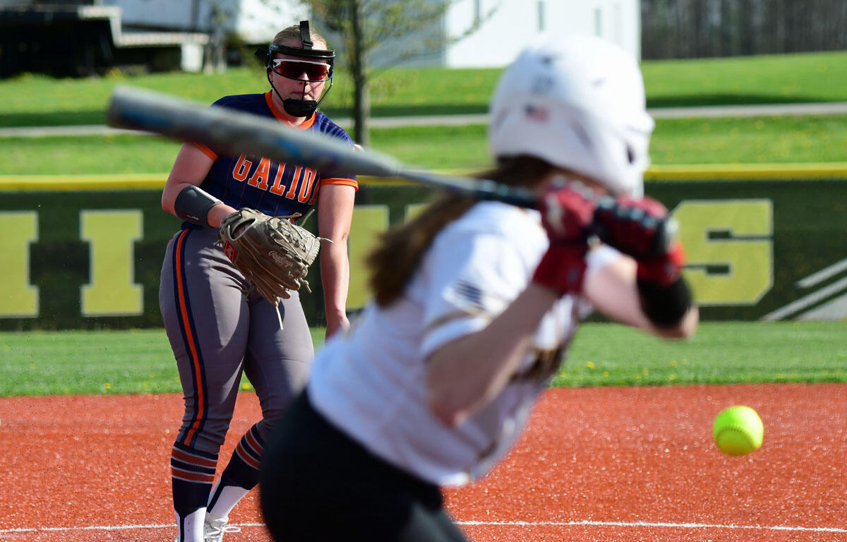 Softball catcher in navy and orange uniform crouches with glove, watching the pitch on a red infield.