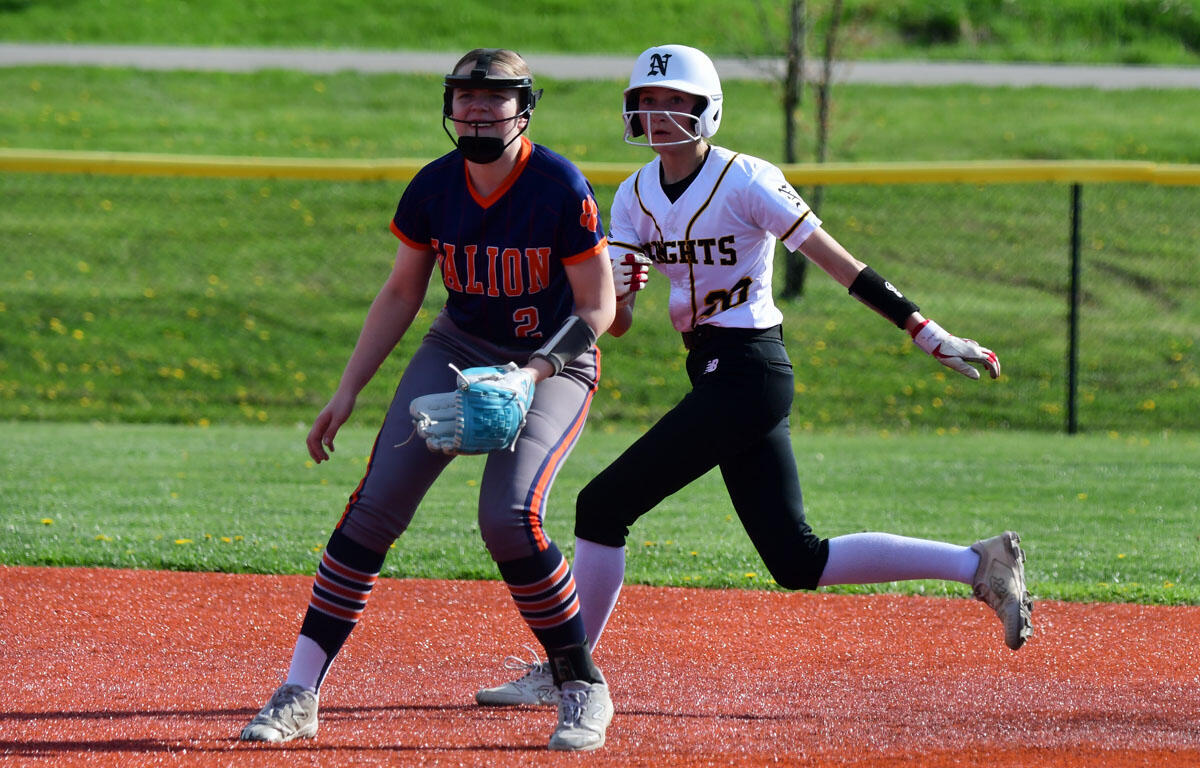 Two female softball players sprint on the infield during a game, one in a dark blue/orange uniform with a glove and helmet, the other in a white jersey with a white helmet and black pants nearby.