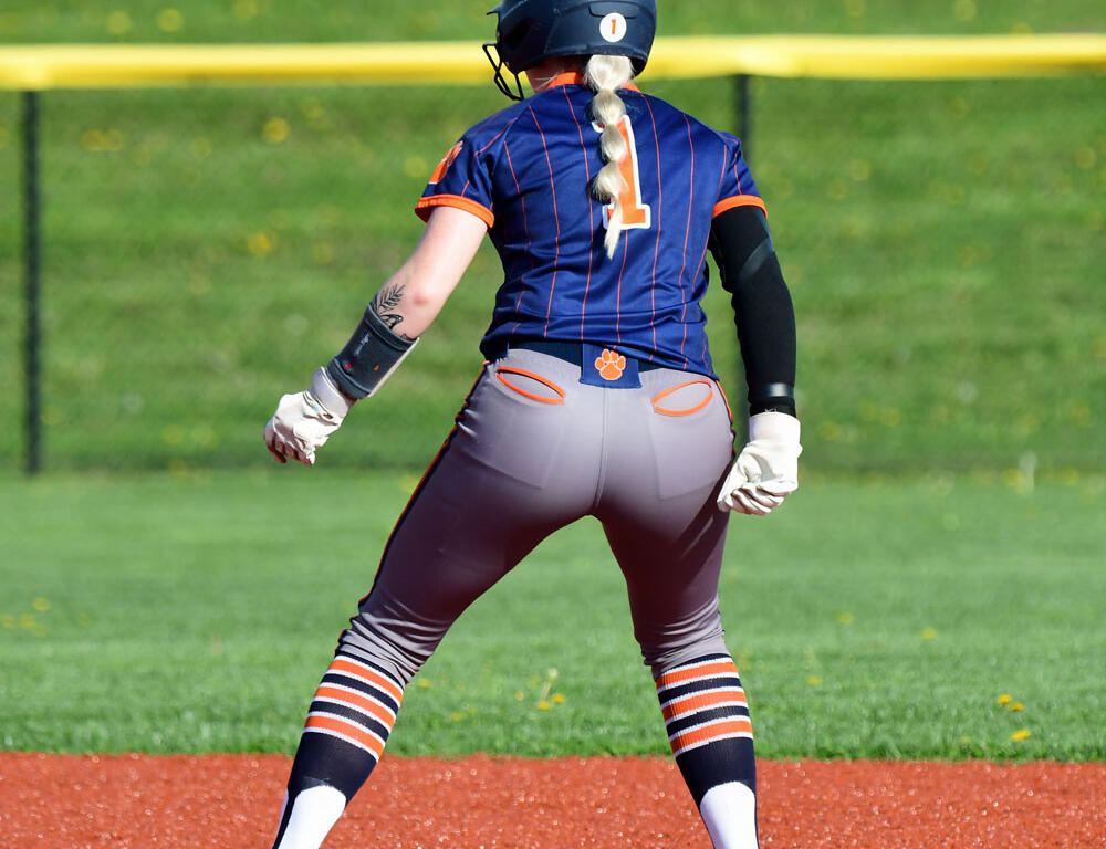 Back view of a female softball player in a blue-orange uniform with number 1, wearing a helmet and gloves on the infield dirt.