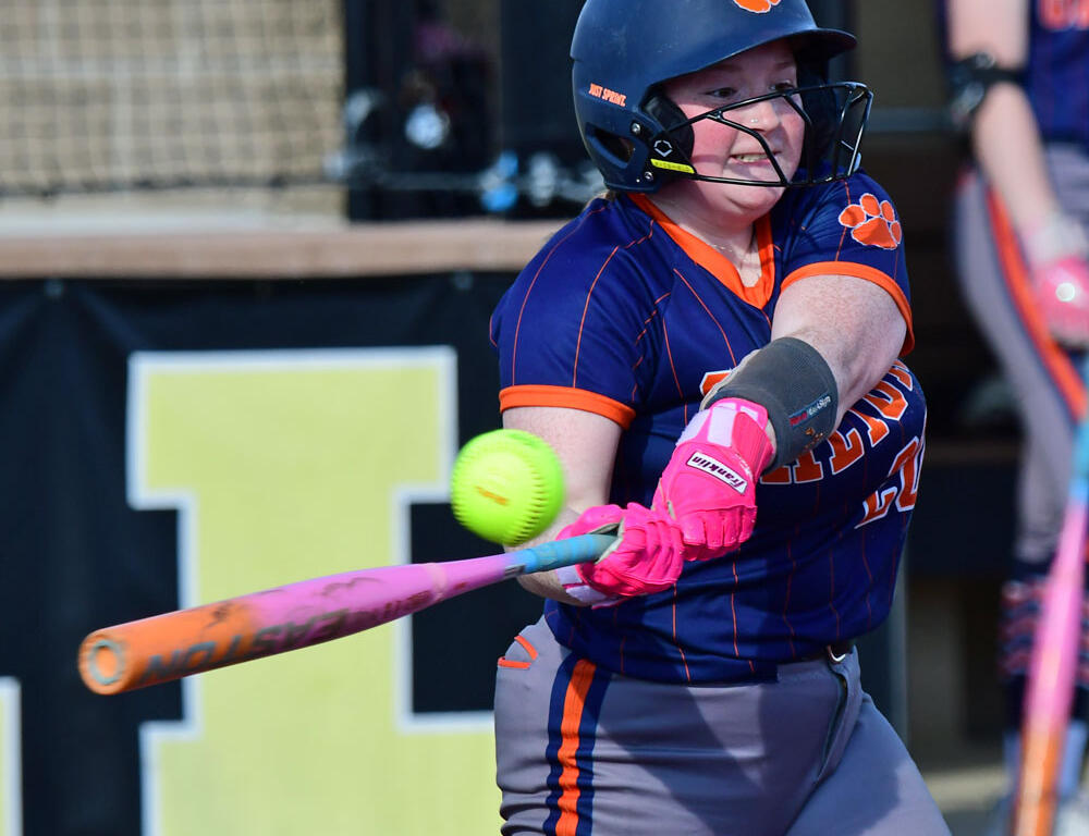 Female softball player in navy and orange uniform swings a pink bat at a bright yellow softball, helmet on, during a game.