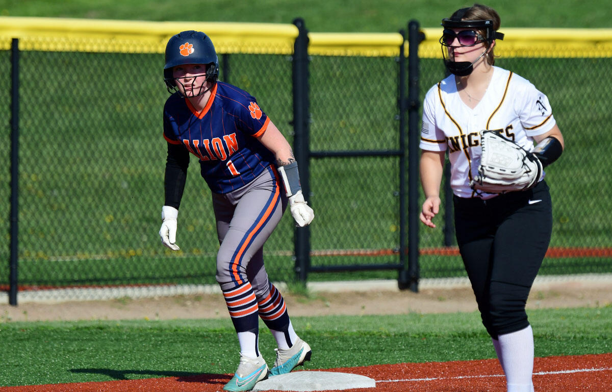 Softball game action: runner in a navy 'LION' jersey slides toward first base in a helmet, with a teammate in white nearby.