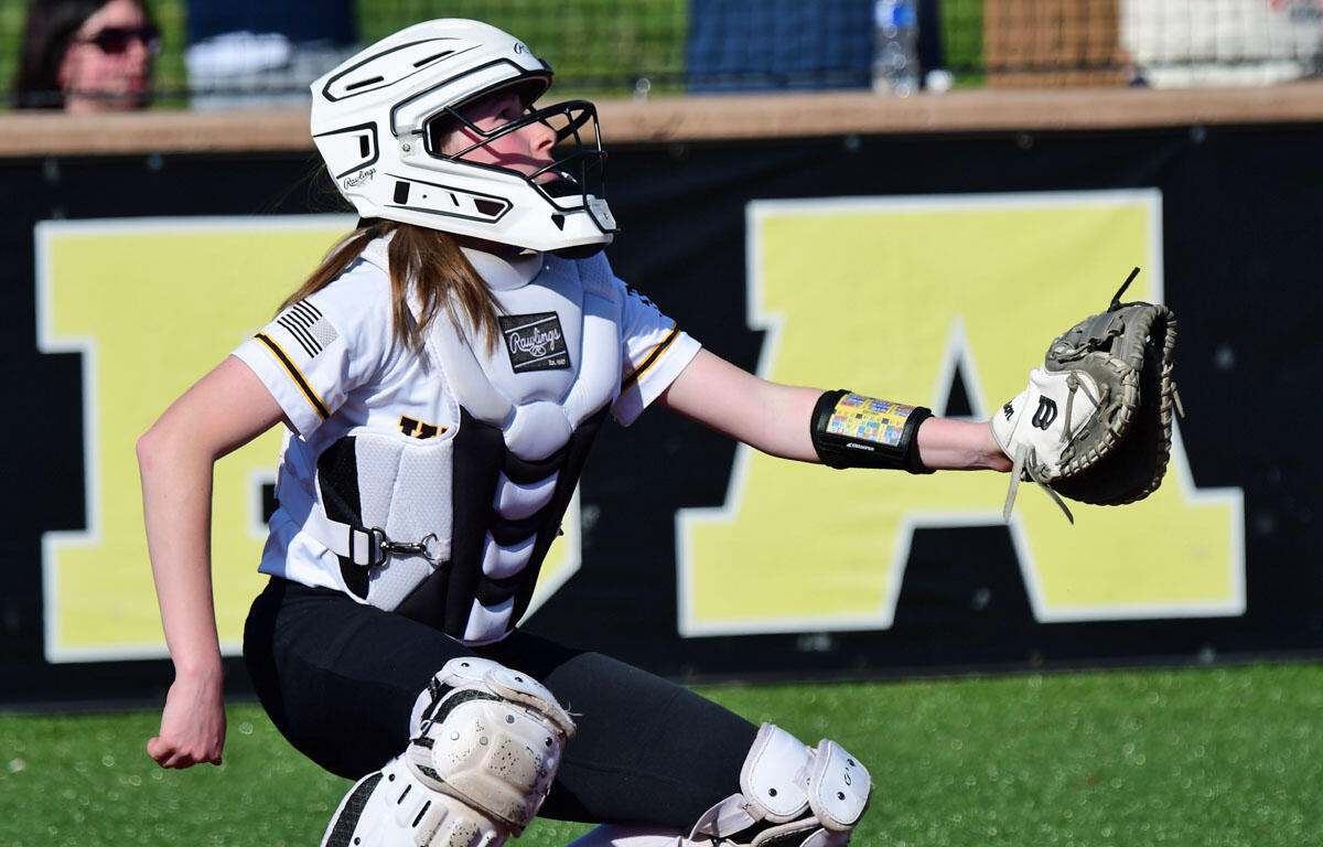 A young catcher in white protective gear and helmet lunges with a glove extended to catch a ball during a game in progress.
