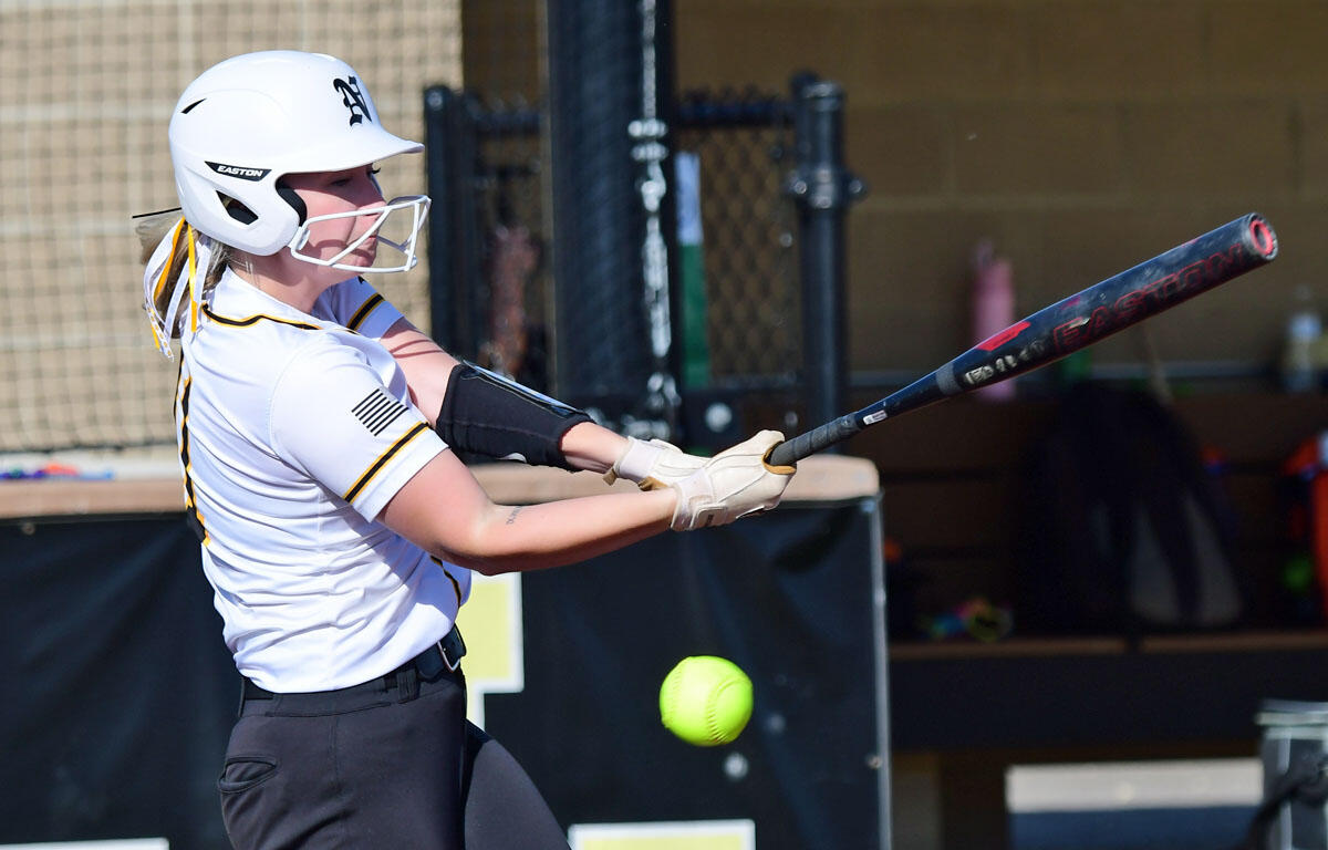 Female softball batter in a white jersey and helmet swings at a pitched neon ball during a game.
