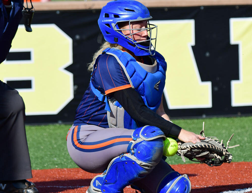 Female softball catcher in blue gear crouching at home plate, glove near a yellow ball ready to field a pitch