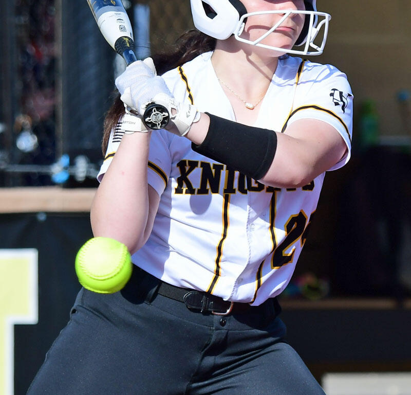 Female softball batter in a white helmet and uniform swinging at a neon-yellow ball during a game, ready to hit.