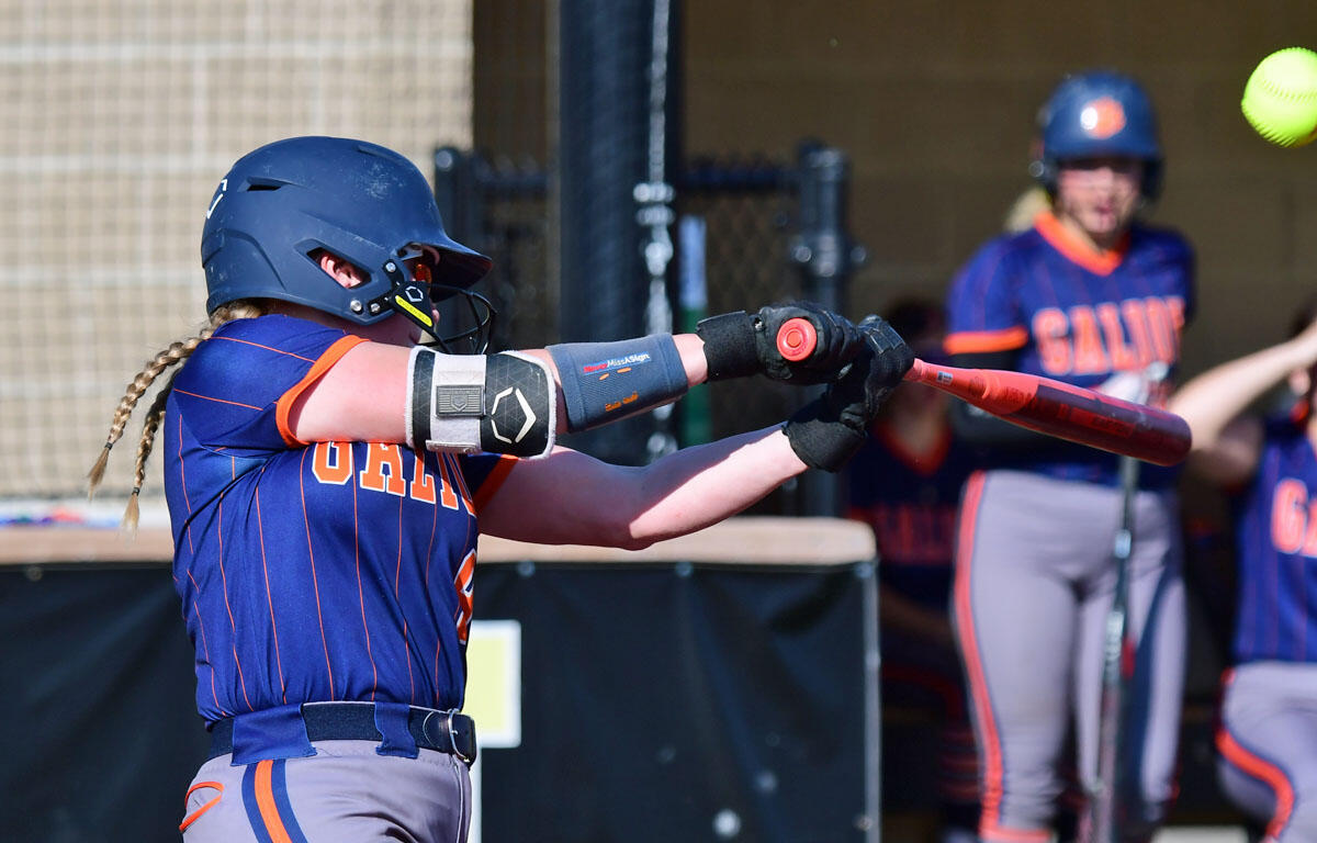 Female softball player in a blue and orange uniform swings a red bat during a game, helmeted and ready at the plate, with teammates in the dugout in the background.