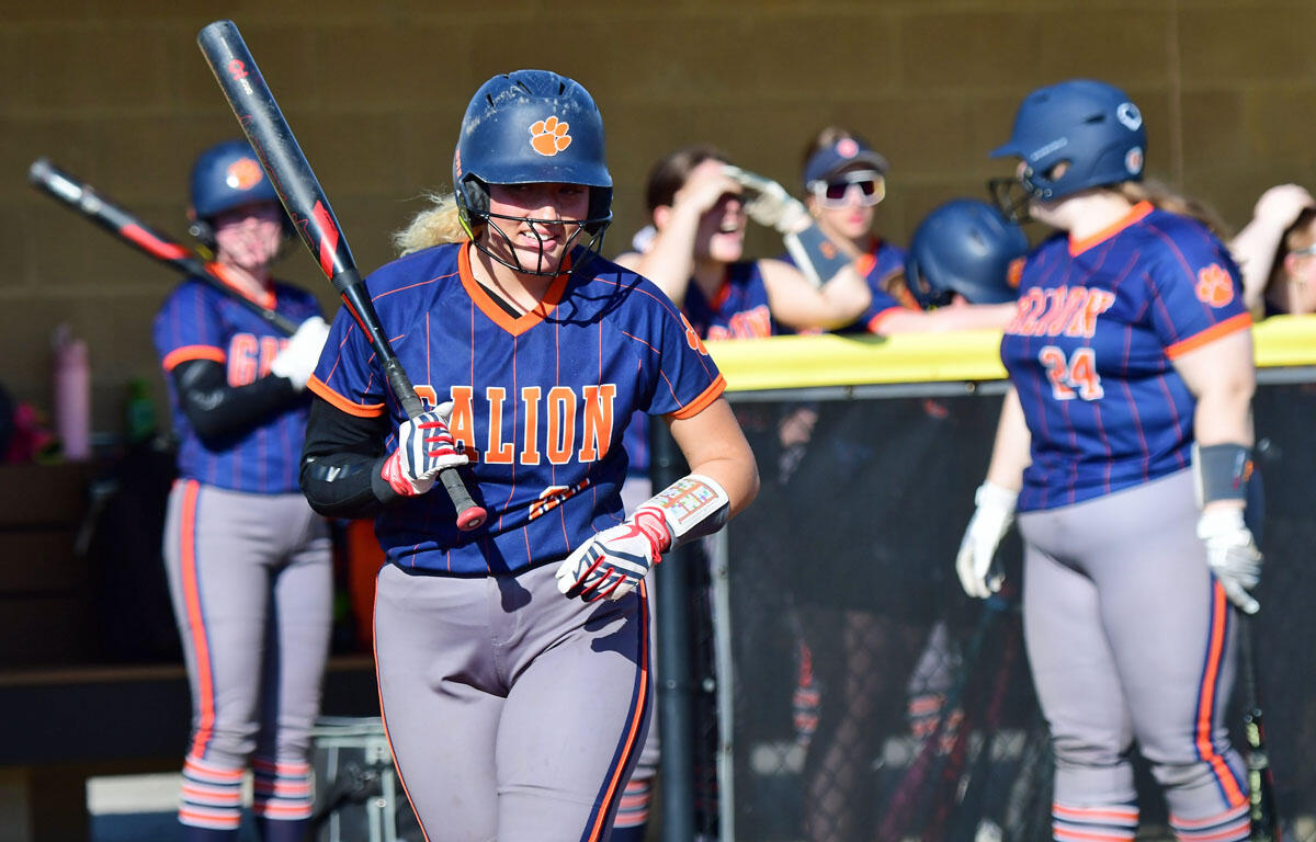 Softball player in blue and orange uniform swings a bat near the dugout as teammates cheer in the background.