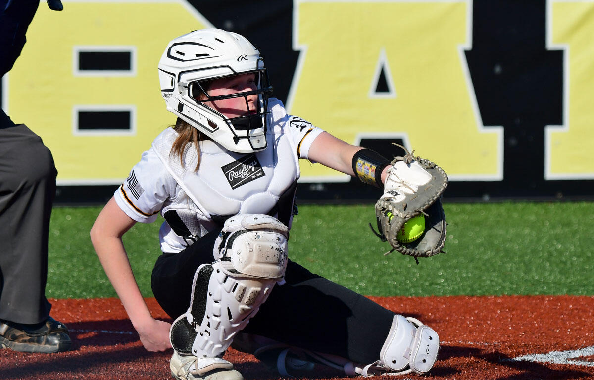 Female softball catcher in white protective gear crouches at home plate, glove ready to catch a bright green ball.