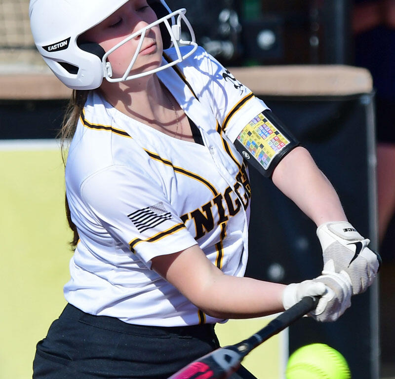 Female softball batter wearing a white helmet and uniform, swinging at a bright yellow softball.