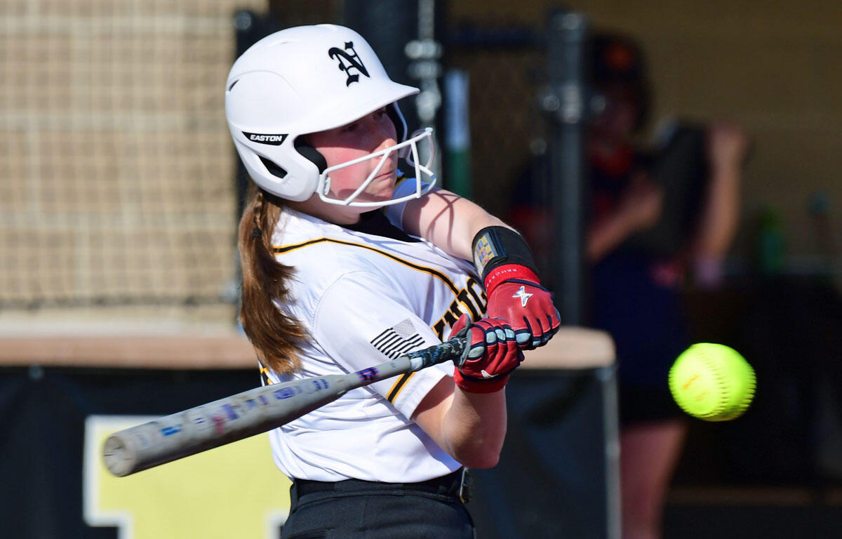 Female softball player in a white uniform and helmet swings at a pitch; neon softball in the air nearby.