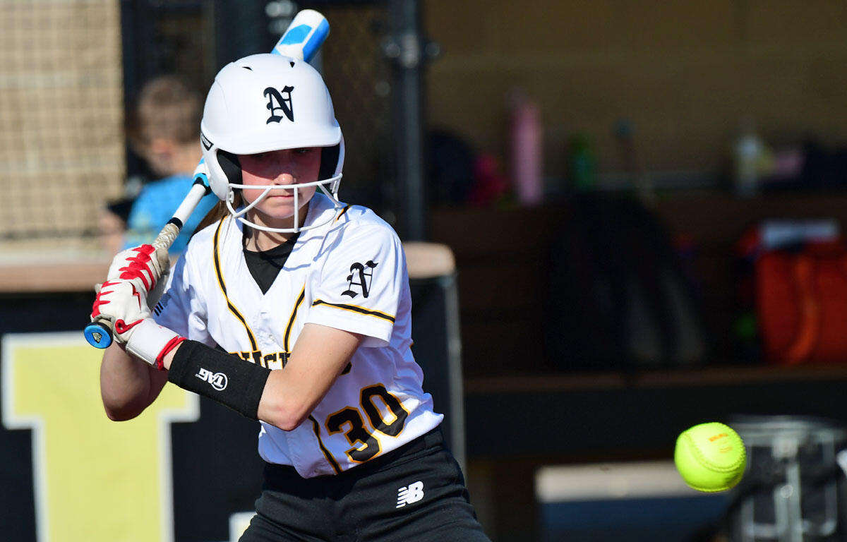 Young softball batter in a white jersey and helmet prepares to swing at a pitched ball near the plate.