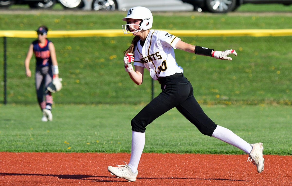 Young female softball player sprinting on a red dirt base path during a game, wearing a white uniform and helmet with a glove in hand.