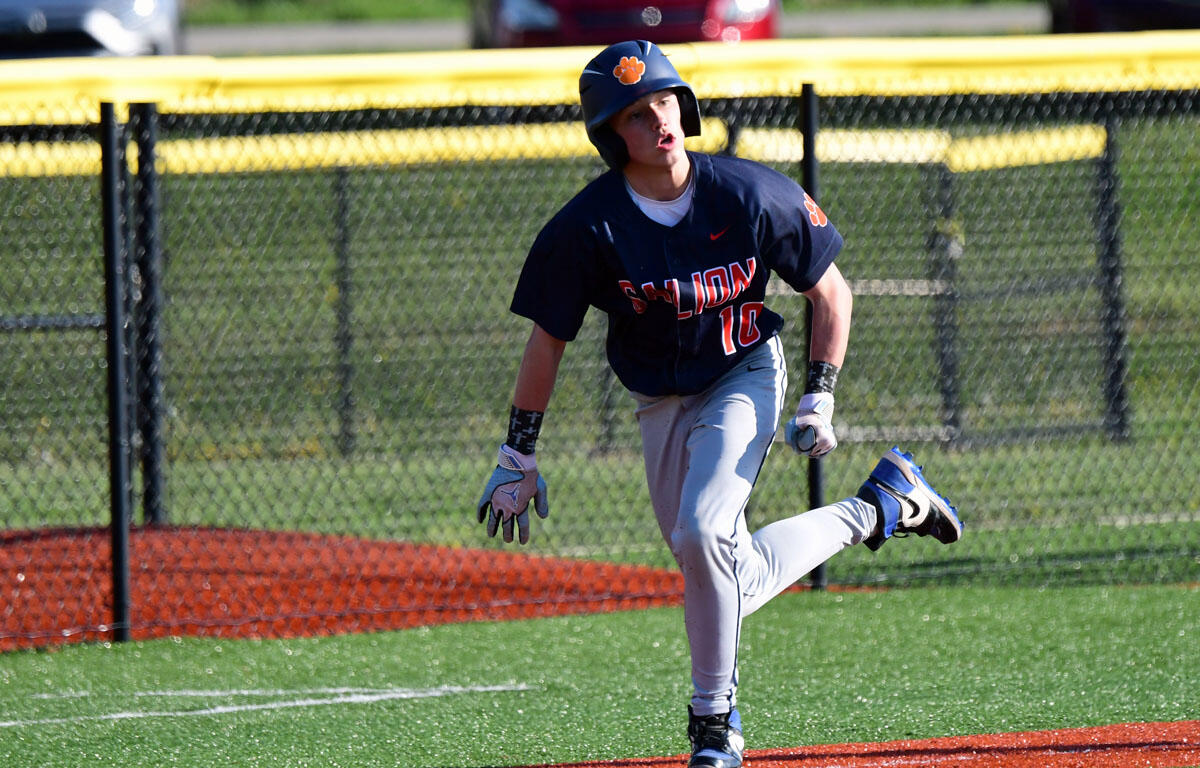 Baseball player in a navy uniform with an orange helmet runs on the field, with a chain-link fence and yellow caution tape in the background.