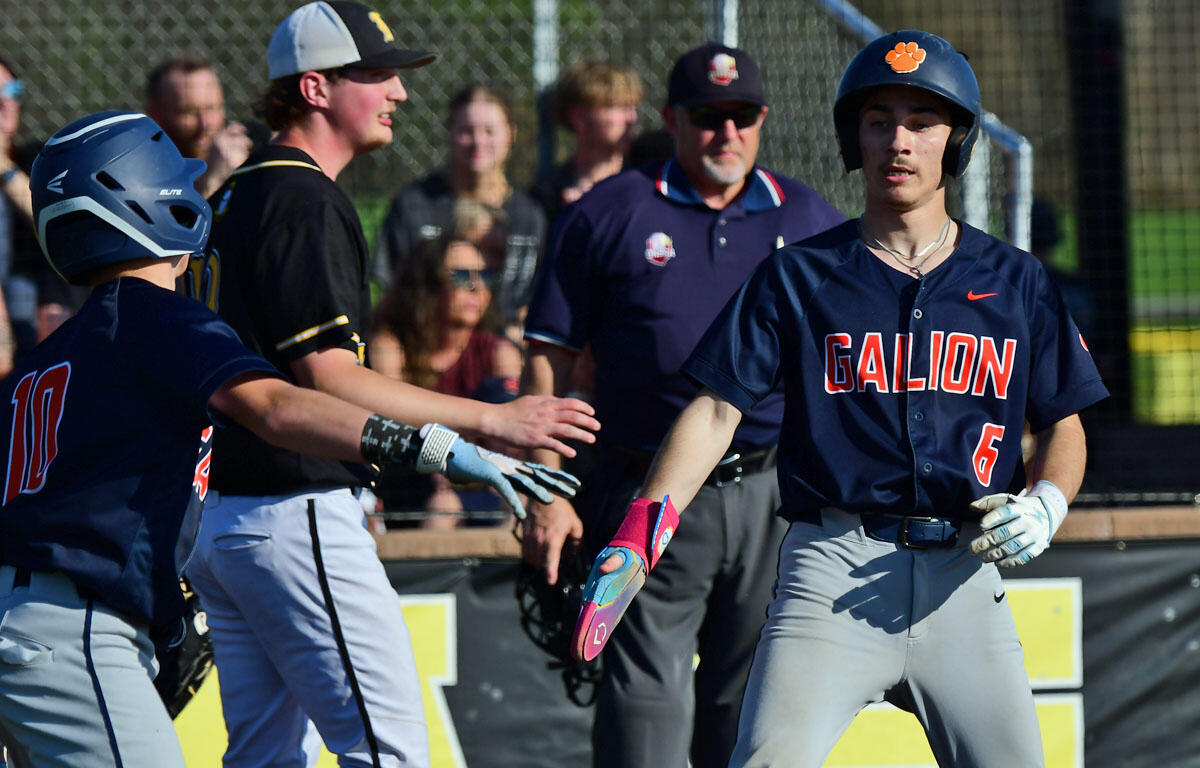 Galion baseball players in navy uniforms exchange a high-five on the field as a coach and fans watch in the background.