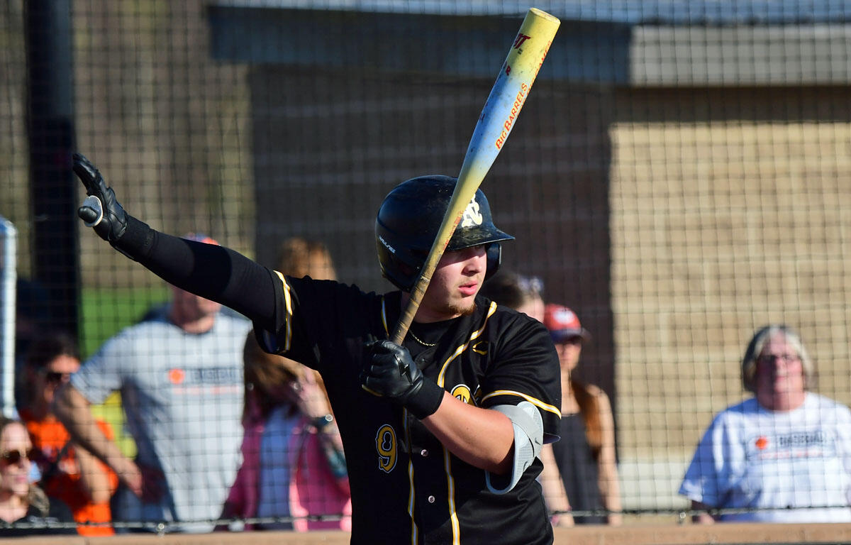 Baseball player in a black and yellow uniform mid-swing with a bat, wearing a helmet as spectators watch behind a chain-link fence.
