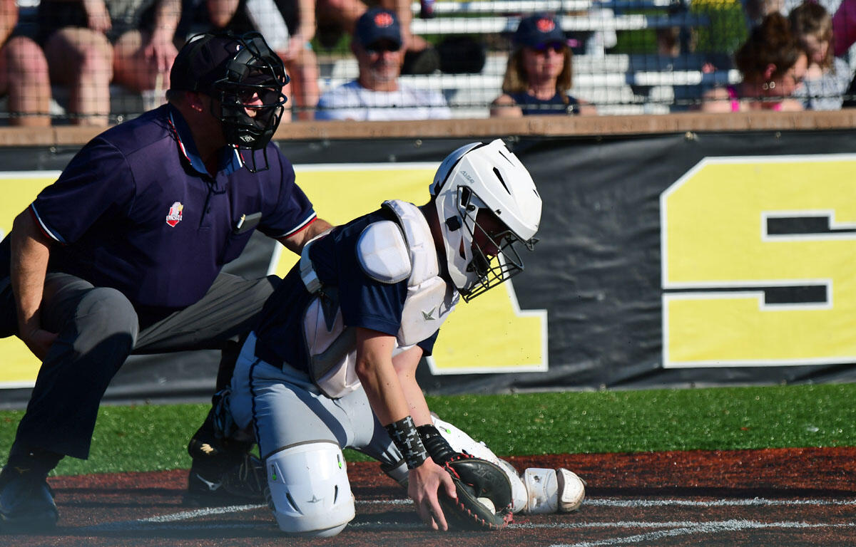Baseball catcher in white gear and navy jersey crouches at home plate, reaching for the ball as the umpire watches behind him.