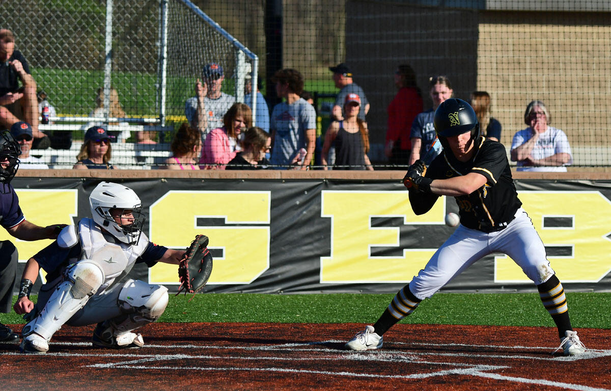 Catcher in white protective gear crouches behind home plate as a batter in black and gold swings at a pitch, with spectators in the background behind a fence.