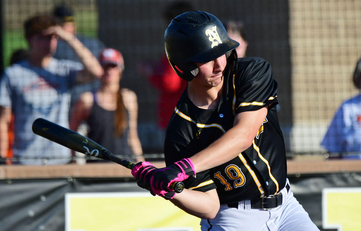 Baseball player in black and yellow uniform swinging a bat, wearing a helmet, on a sunny field with fans in the background, mid-swing.