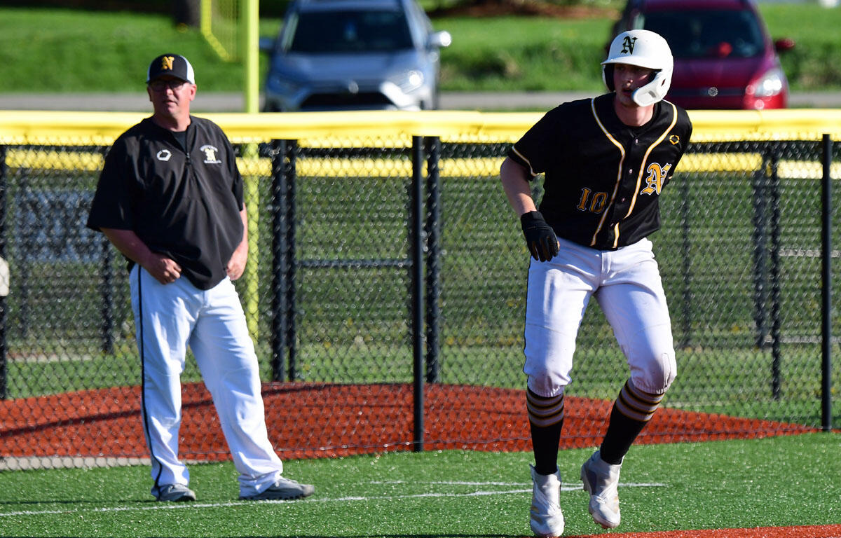Baseball coach in black shirt and white pants watches a runner in a black and gold uniform sprinting on the artificial turf near a chain-link fence and yellow outfield wall.