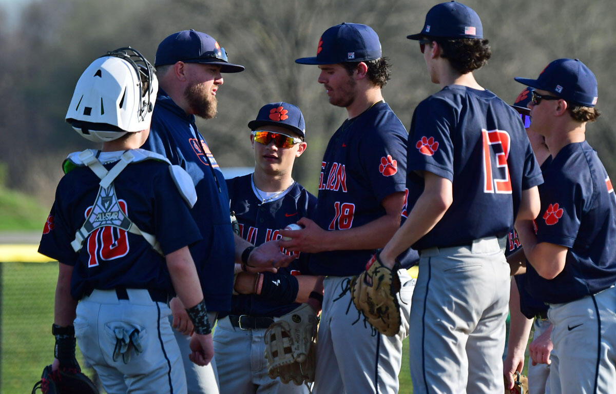Baseball team huddle on the field with coach speaking to players in navy uniforms and a catcher in gear on the left.