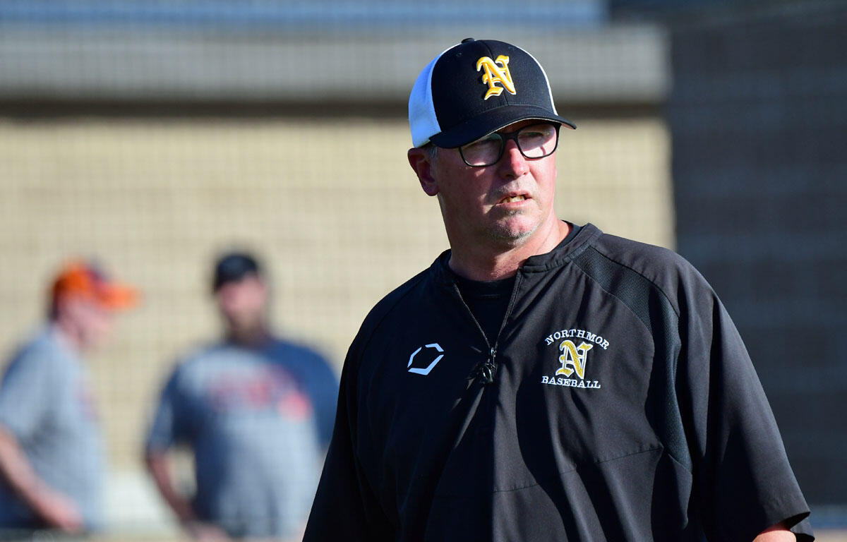Baseball coach wearing a black Northmor jacket and cap, standing on a field with blurred players in the background