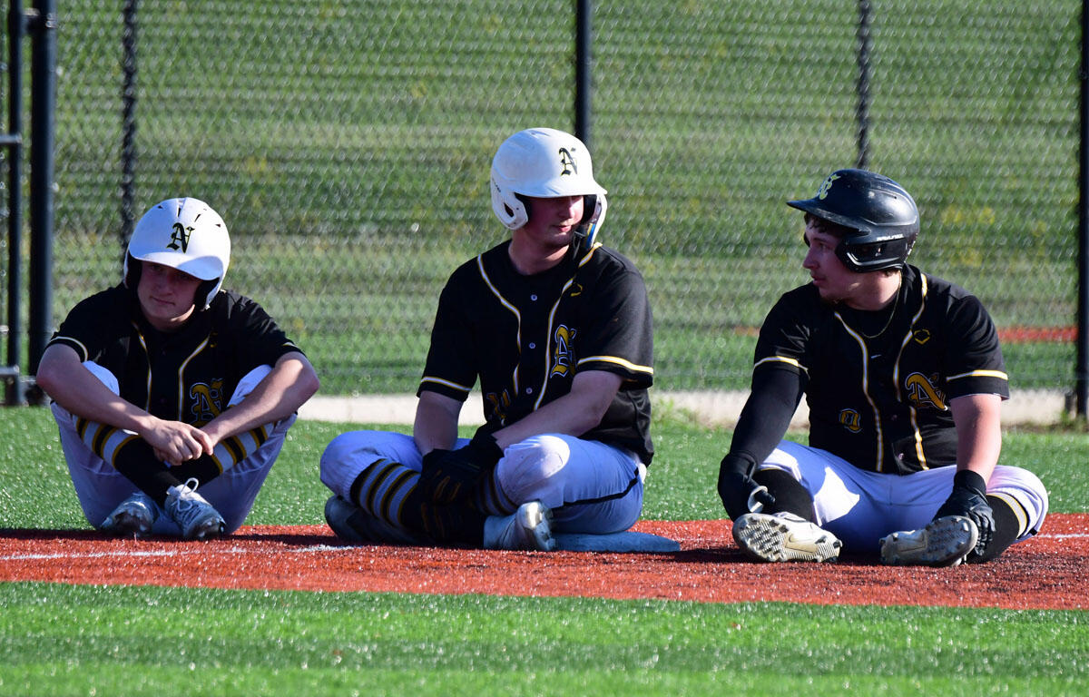 Three baseball players sit on the infield grass near the dugout, wearing black and gold uniforms and helmets.