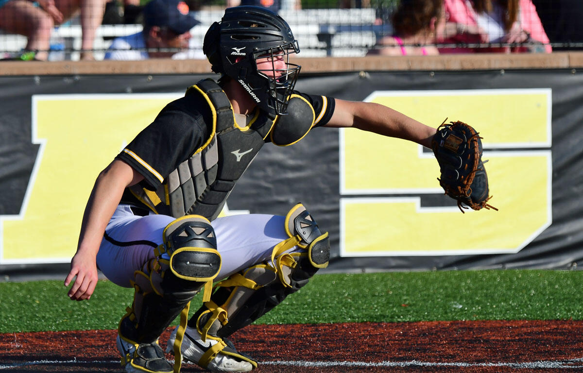 Baseball catcher in black and yellow protective gear crouches with glove extended to catch a pitch during a game to the left of home plate.