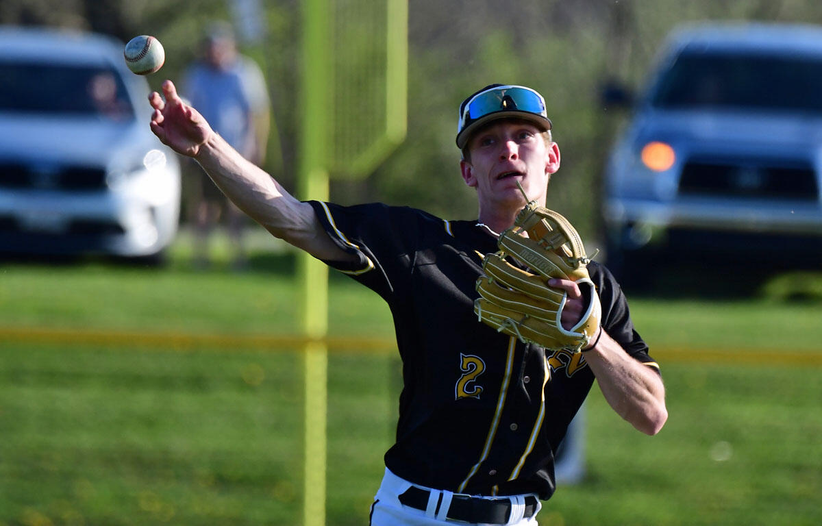 Right-handed baseball pitcher in a black and gold jersey delivers a throw on a sunny field with his glove hand up front.