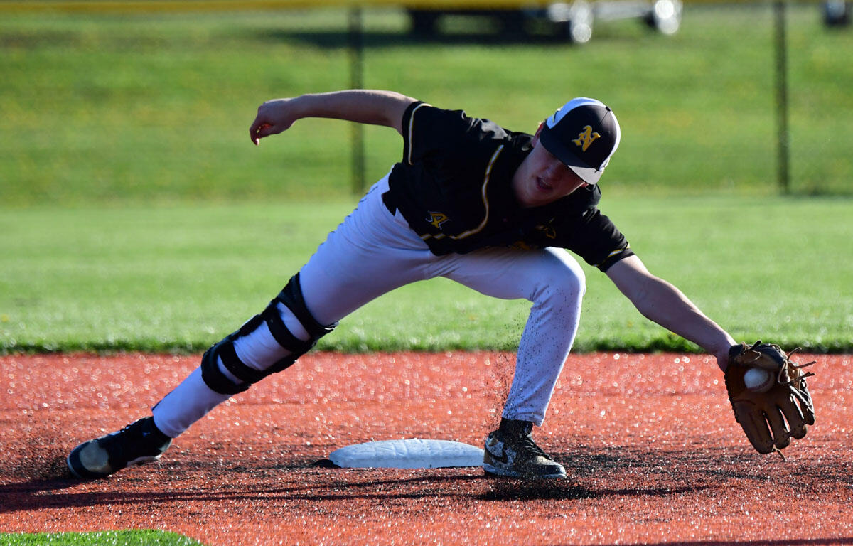 Baseball infielder slides into a base, glove reaching for the ball on a red dirt infield while teammates watch in the background.