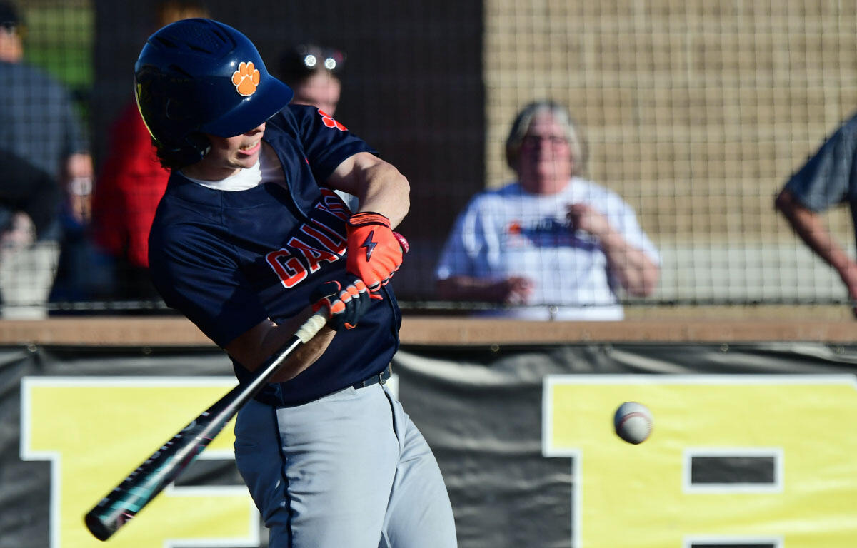 Baseball player in a navy uniform swings at a pitch, helmet with an orange pawprint, ball in midair nearby behind the bat.