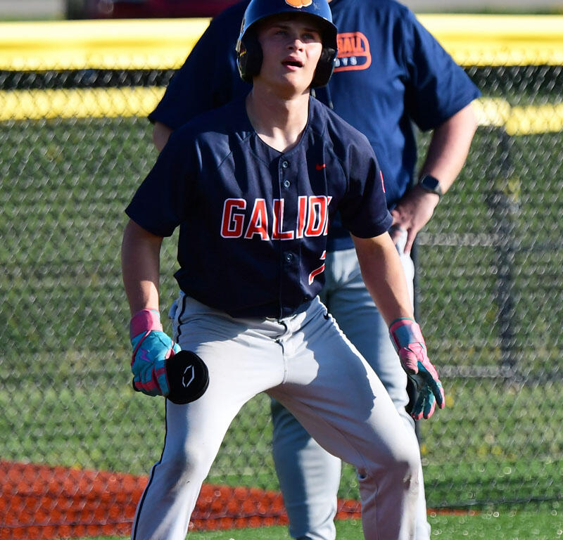 Young baseball player in navy uniform with 'GALIO' on the chest, wearing a helmet and batting gloves, crouched in a ready stance on a bright field.