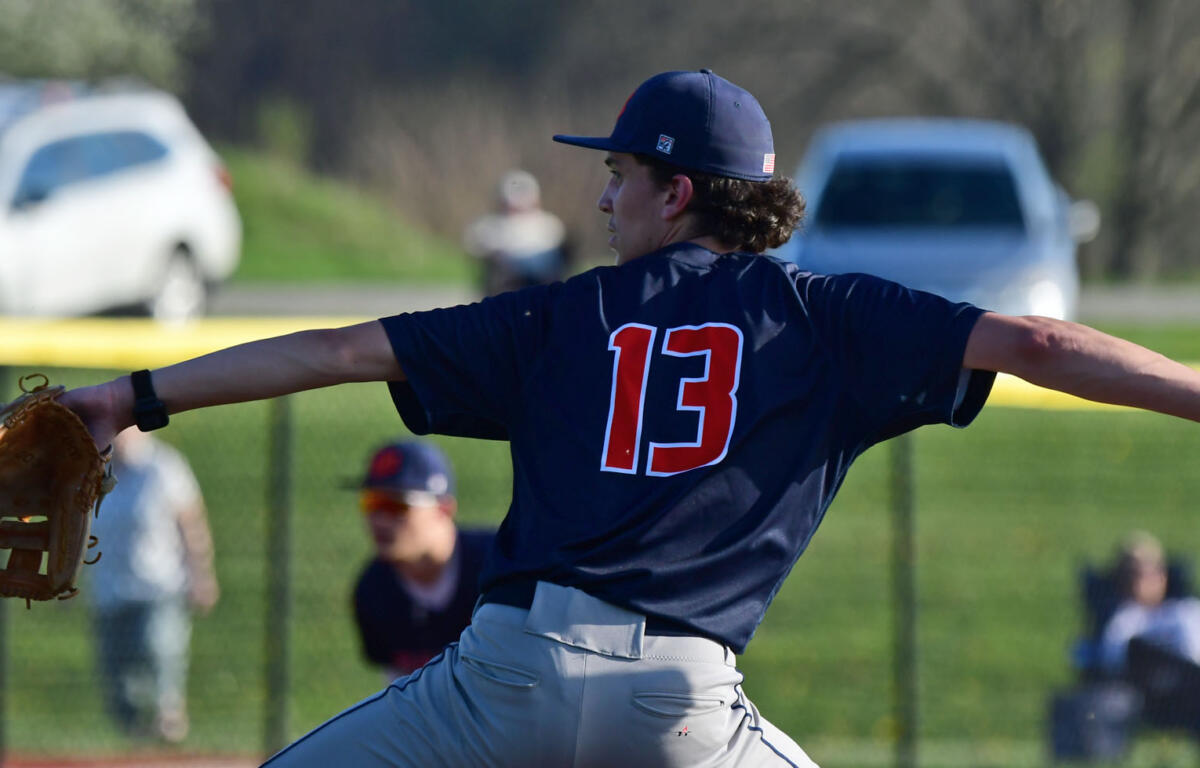 Baseball pitcher in a navy jersey with number 13 winds up to throw a ball on a sunny field, glove on left hand.