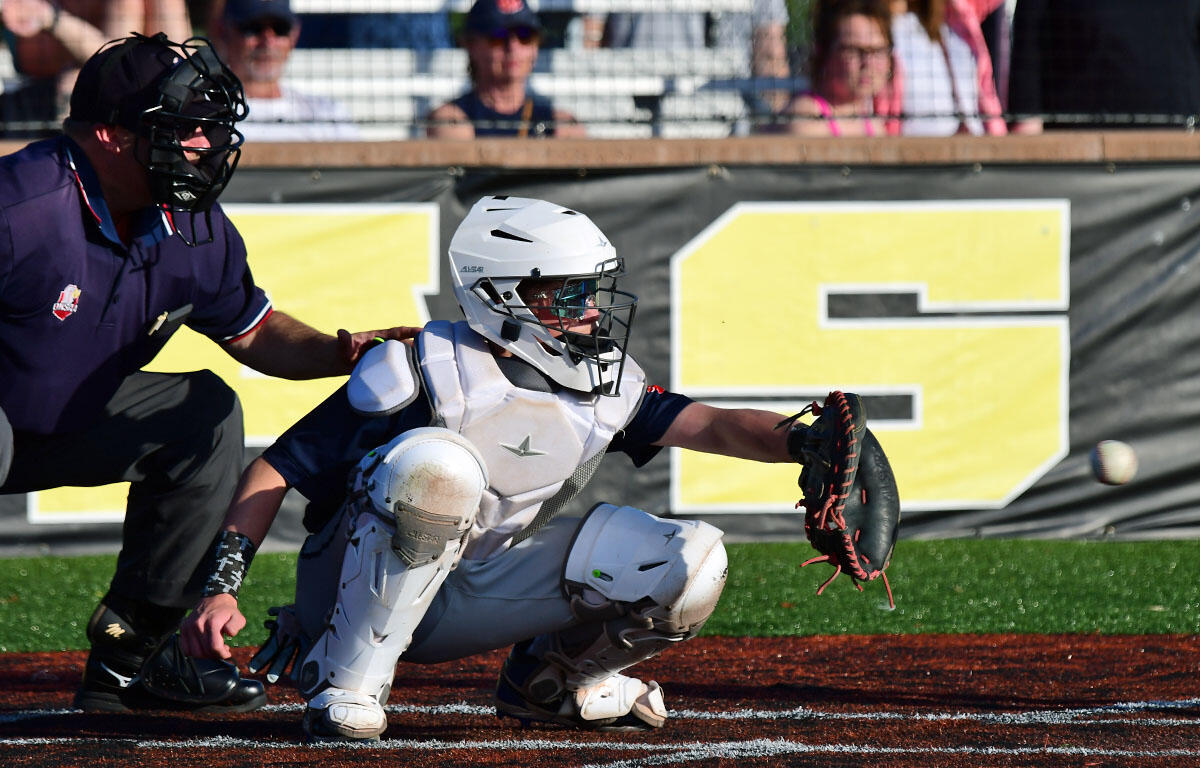 Baseball catcher in white protective gear lunges to catch a ball at home plate as an umpire watches from behind the action center fielded by a dark-uniformed umpire.