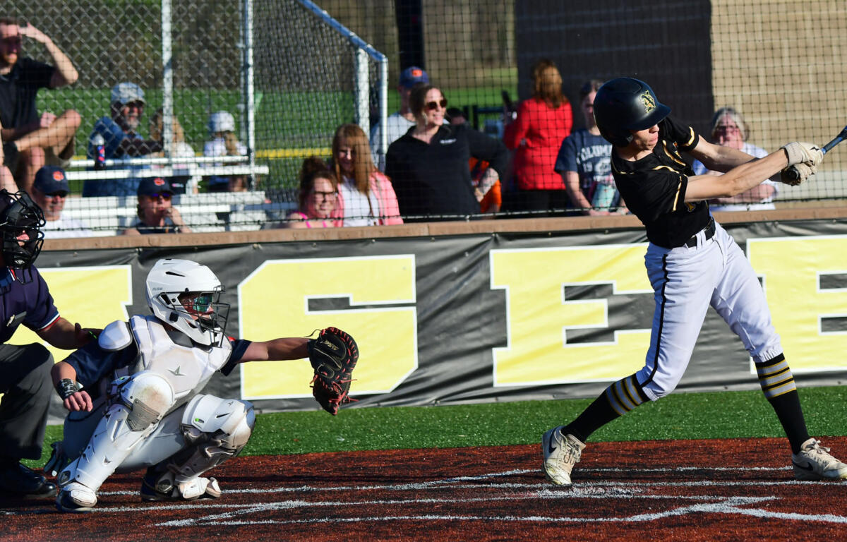Baseball batter in black and white swings at a ball as catcher and umpire guard home plate and spectators watch from behind the fence.