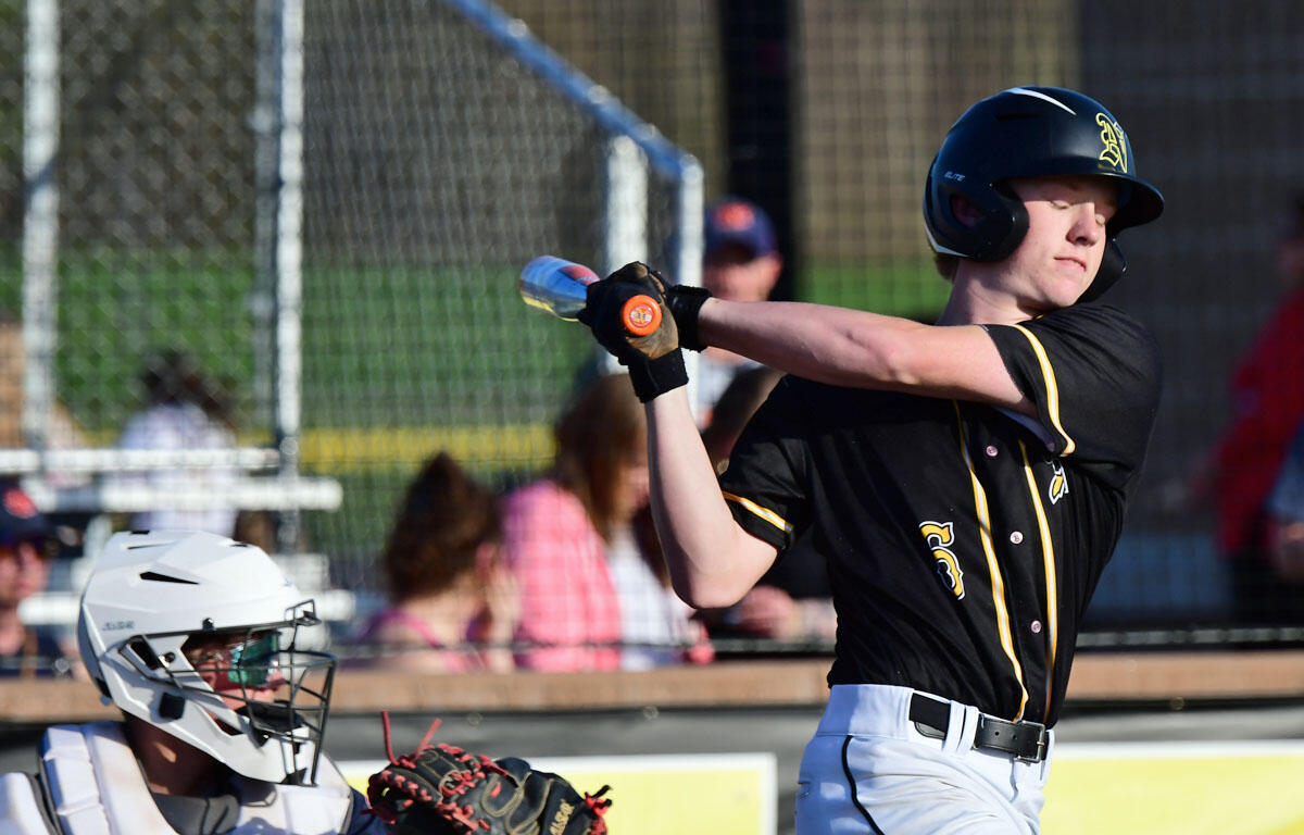 Baseball batter in a black and yellow uniform swings at a pitch, with a white-gear catcher behind him in a fenced field.