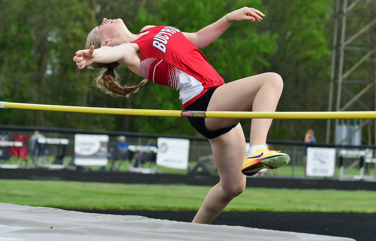 Female high jumper in a red uniform clears a yellow high jump bar outdoors. She arches mid-air with arms outstretched.