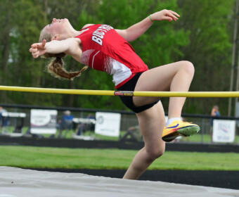 Female high jumper in a red uniform clears a yellow high jump bar outdoors. She arches mid-air with arms outstretched.