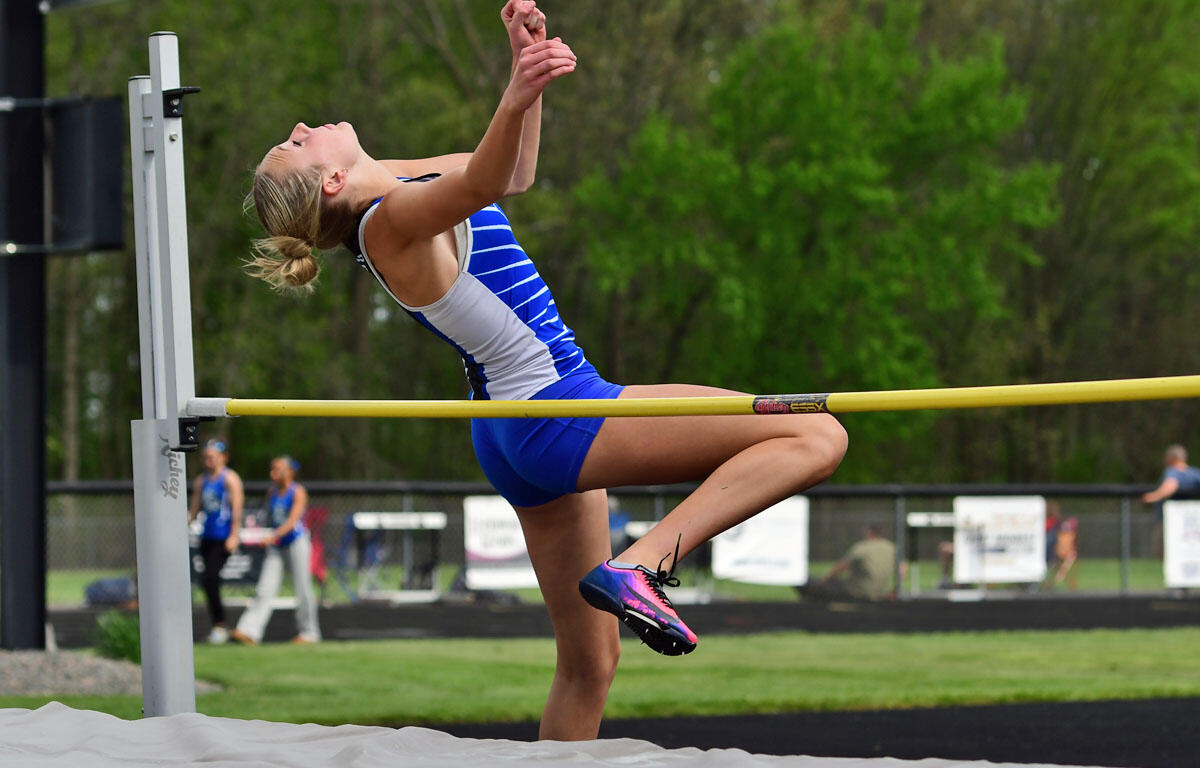 Female athlete in a blue and white uniform clears a high jump bar outdoors during a track meet.