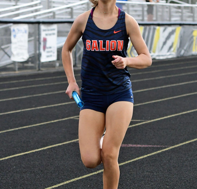 Female sprinter in a navy Galion uniform runs on a track holding a blue baton.