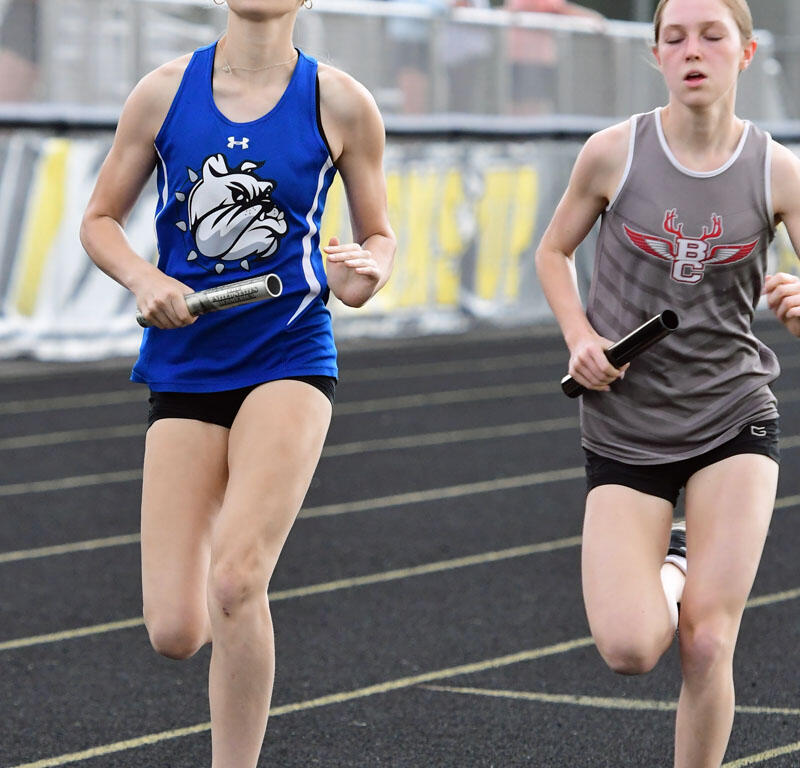 Two young girls run a relay on a track, each holding a baton with spectators behind them.