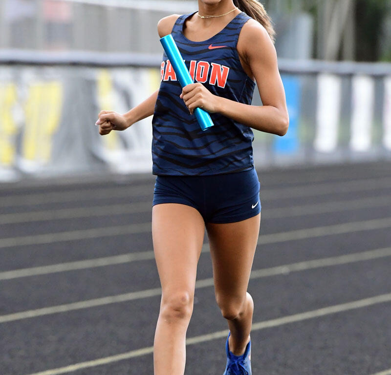 Female sprinter in a blue athletic uniform running on a track, holding a blue relay baton mid-race.
