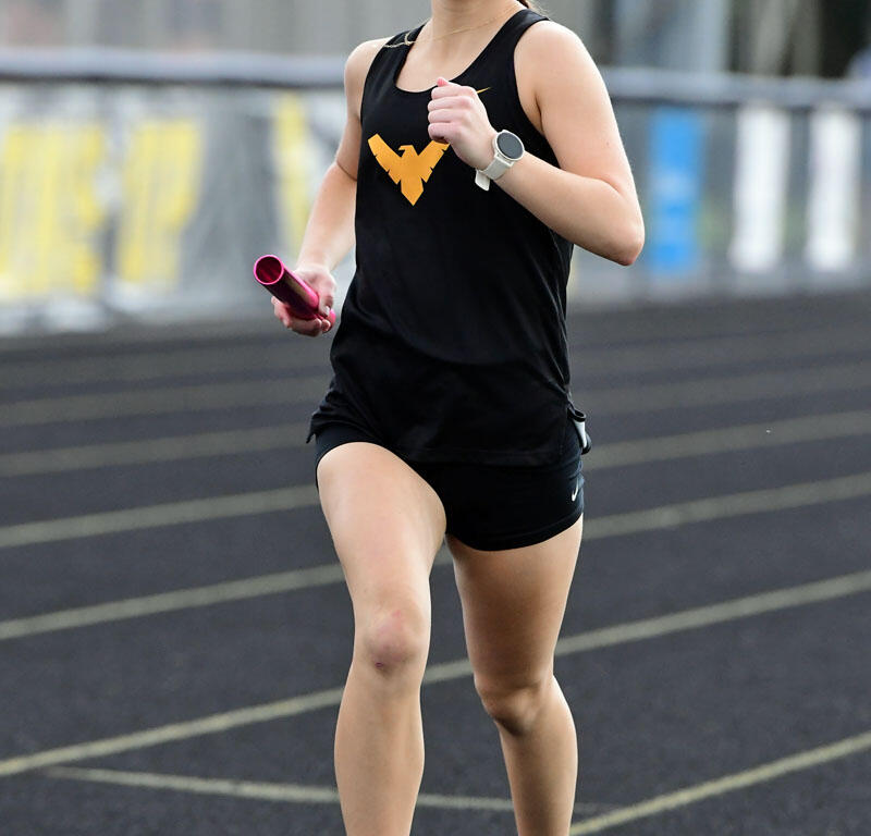 Young female runner in a black tank top and shorts, sprinting on a track while holding a pink baton and wearing a watch, focused on the race distance la track lane markers nearby.