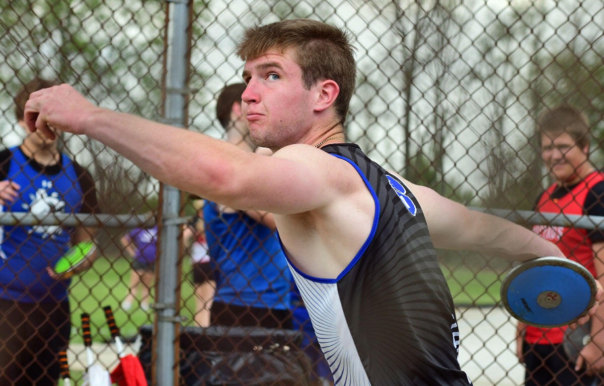 Athlete in a black sleeveless jersey throws a blue discus, extending his arm with a chain-link fence behind him and spectators nearby.