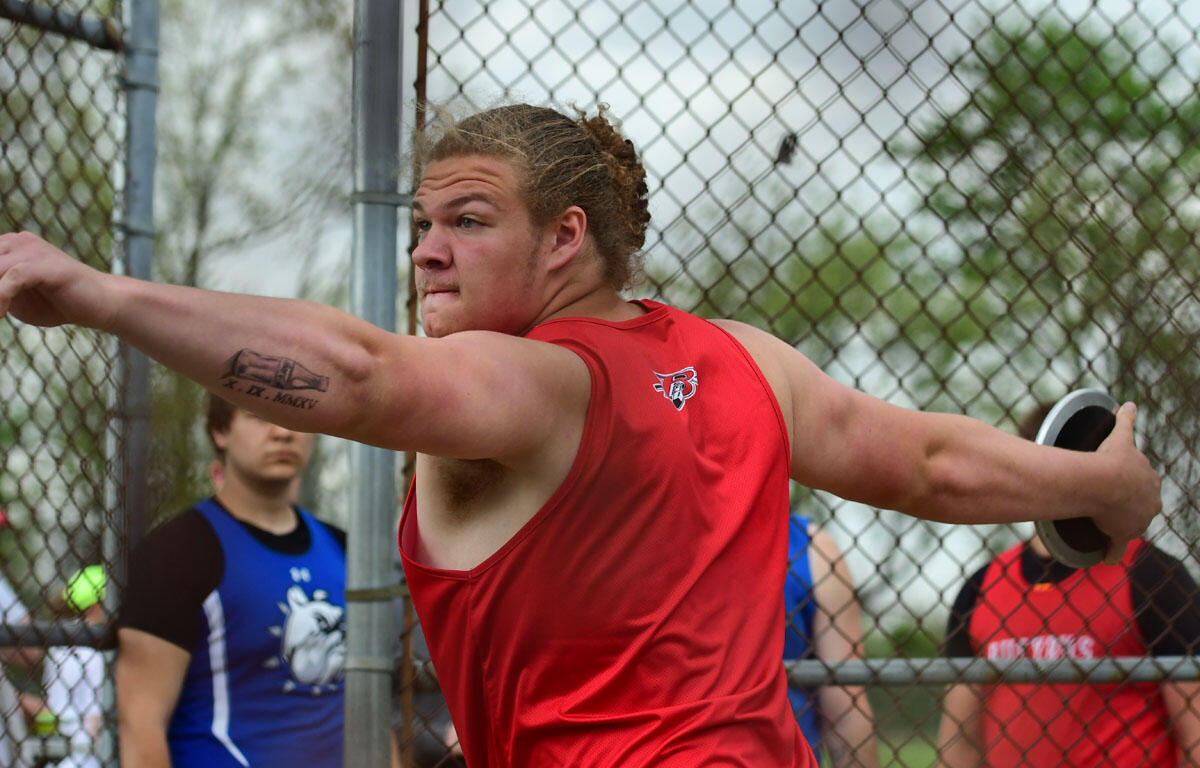 Athlete in a red singlet throws a discus during a track and field event behind a chain-link fence.