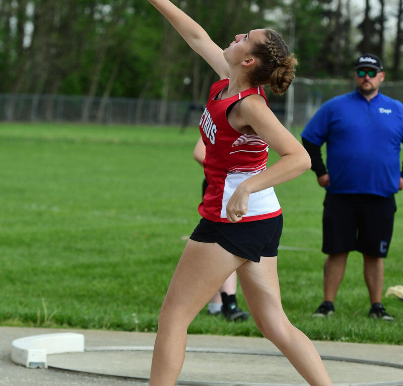 Female athlete in red uniform mid-throw of a shot put on a track and field circle, with a coach watching in the background.