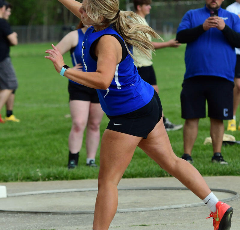 Female hammer thrower in a blue jersey and black shorts powers the hammer overhead during a track-and-field event, with onlookers in the background.