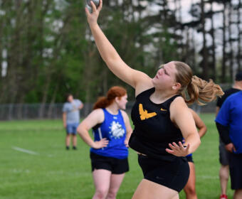 Female athlete in black uniform releases a shot put from the throwing circle on a grassy field, neon green shoes visible.
