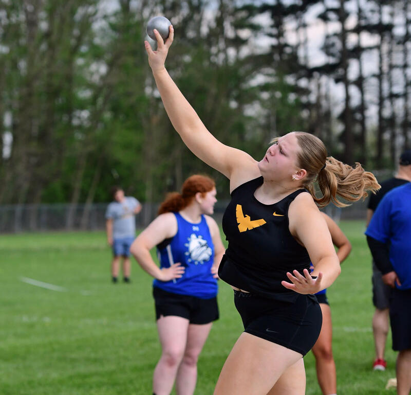 Female athlete in black uniform releases a shot put from the throwing circle on a grassy field, neon green shoes visible.